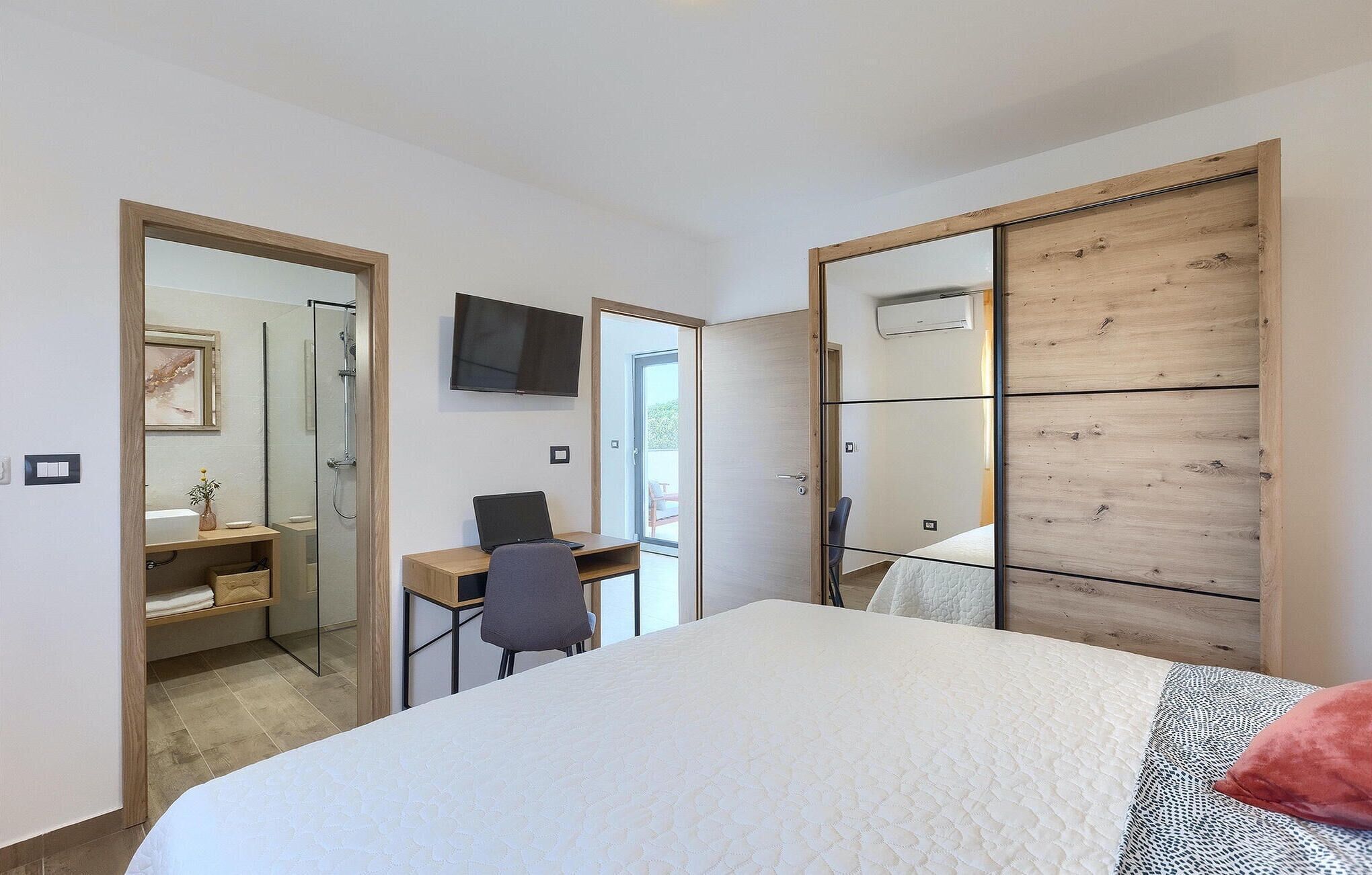 A bedroom with a bed, a desk with a laptop, a chair, a TV, and a sliding door leading to a bathroom.