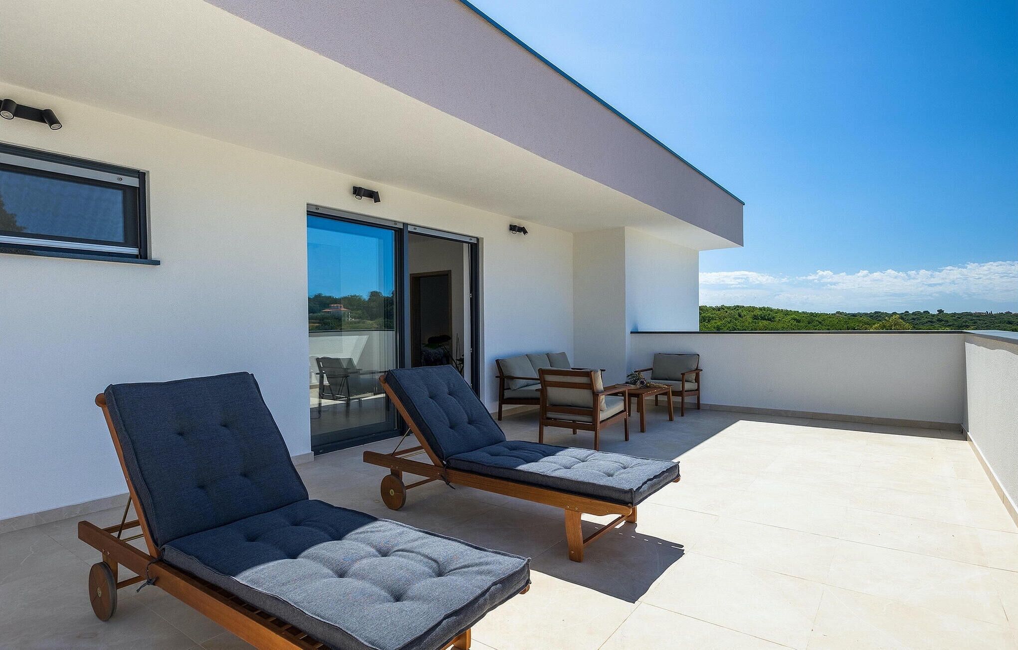 A modern outdoor seating area with two lounge chairs and a table, a glass door leading to an interior space, and a clear view of greenery in the background.