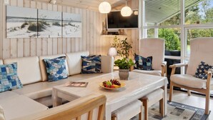 Un salón acogedor con un sofá blanco, una mesa de comedor y dos sillas. Hay dos televisores instalados en la pared, una planta en maceta y un frutero sobre la mesa.