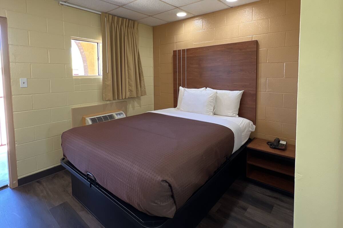A hotel room with a single bed, a wooden headboard, a small bedside table, a telephone, and a window with curtains.