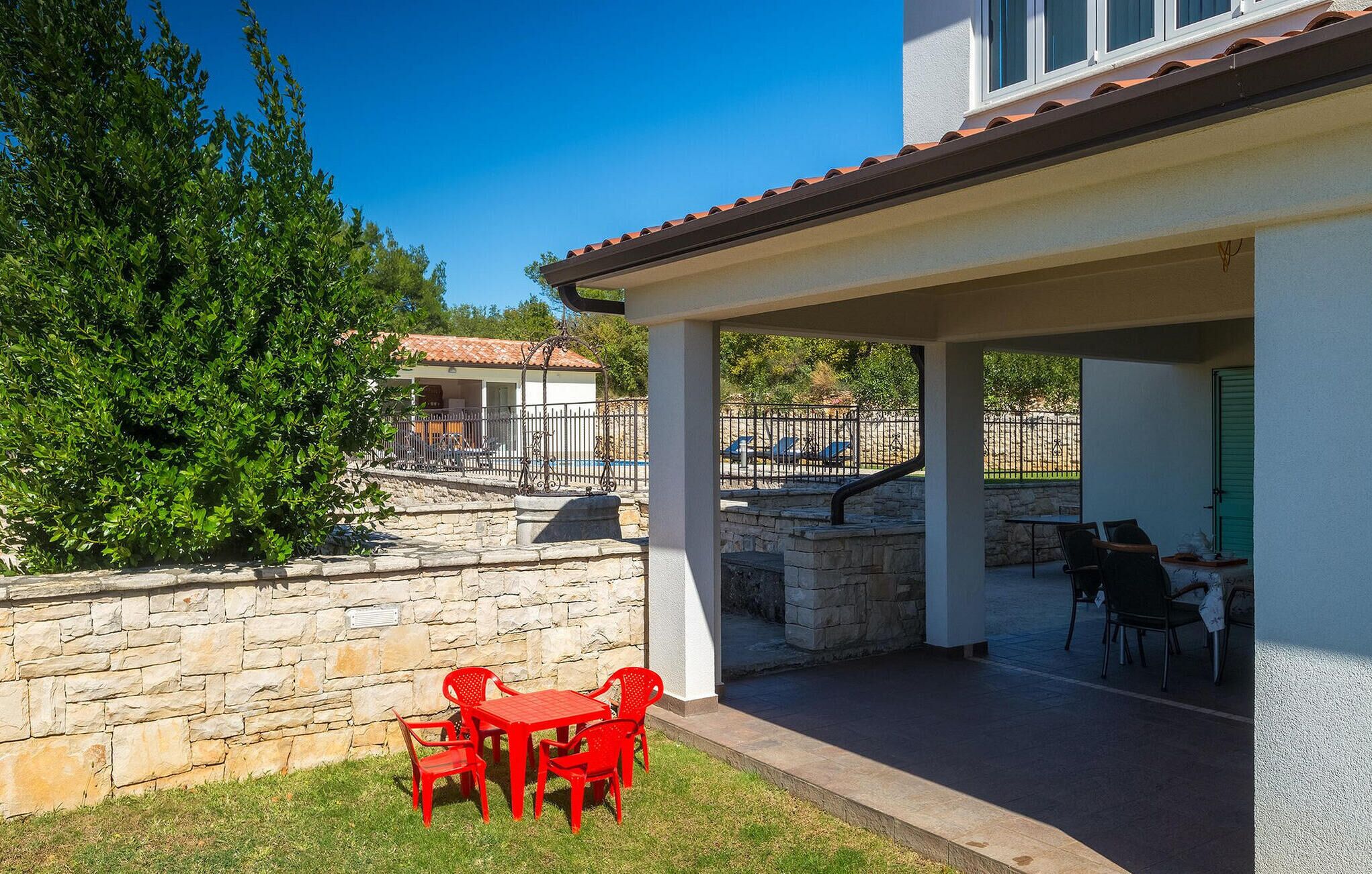 Uma área de varanda coberta com uma parede de pedra, uma mesa com cadeiras vermelhas e uma vista para uma piscina e espreguiçadeiras ao fundo.