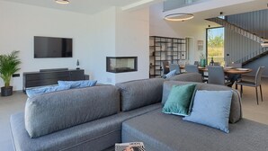 Un salon moderne comprenant un canapé d’angle, un téléviseur à écran plat, un coin repas avec une table en bois et un escalier menant à un niveau supérieur.
