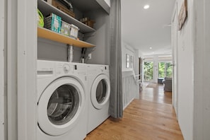 Une buanderie équipée de deux machines à laver à chargement frontal, d’une étagère contenant des produits de nettoyage et d’un plancher en bois.