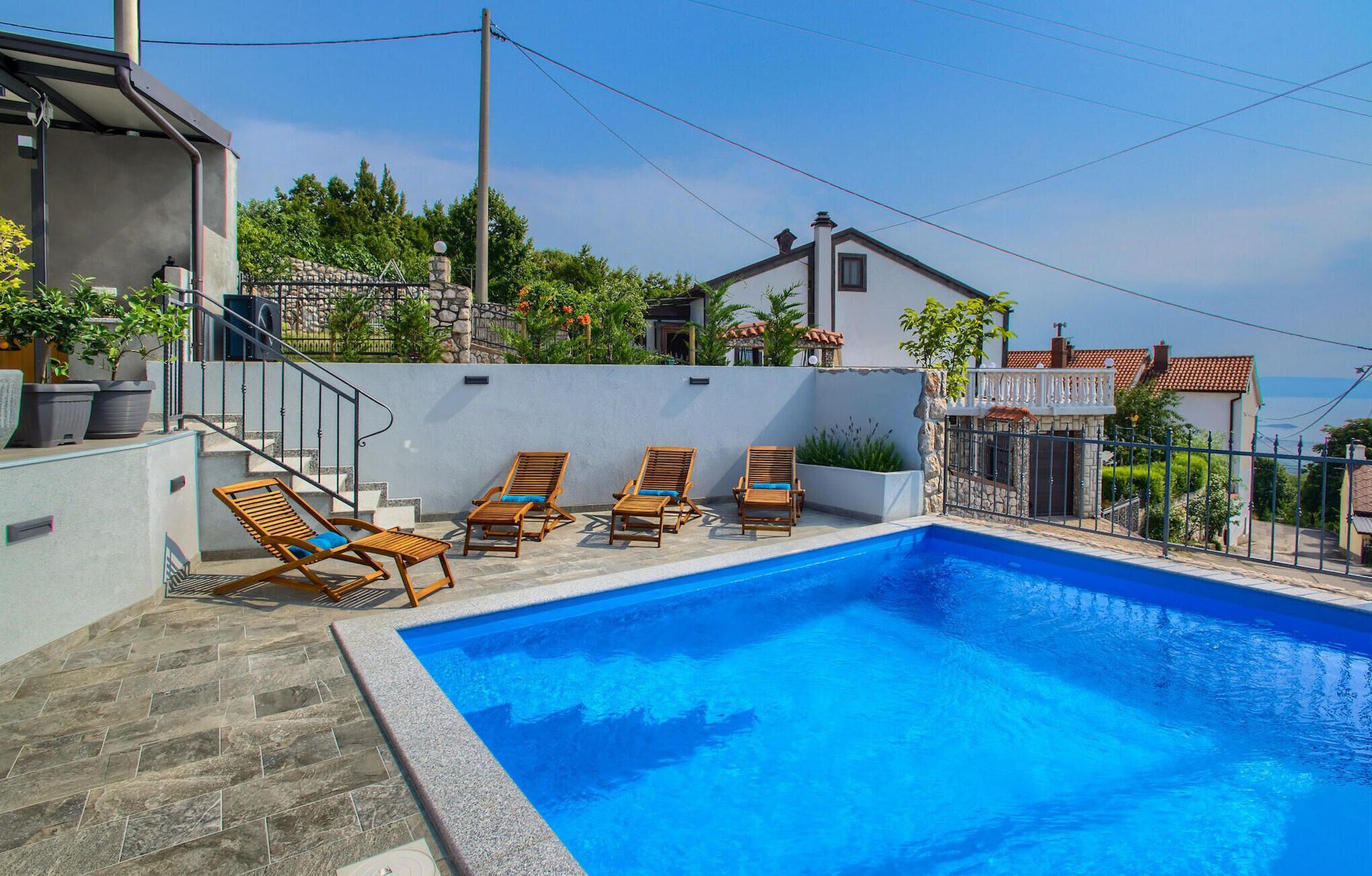 A swimming pool with four wooden lounge chairs, a stone wall, and a view of neighboring houses.