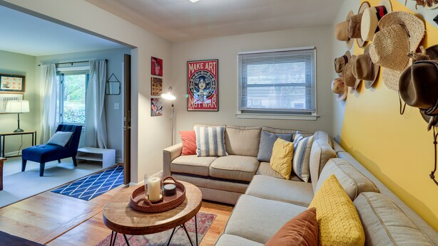 A living room with a sofa, a coffee table, and a wall decorated with hats. There is a window with blinds and a door leading to another room. - Ann Arbor Home w/ Deck, 2 Mi to Michigan Stadium! (Ann Arbor)