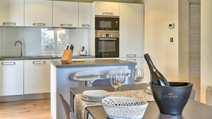 A modern kitchen with white cabinets, a built-in oven, and a microwave. There is a dining area with a table set for two, a bottle of champagne, and a basket. - Awesome home in Opatija (Opatija)
