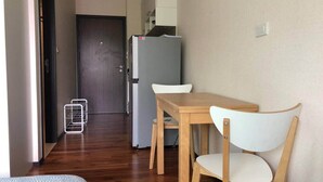 A small, compact room with a wooden table and a chair, a bed, a kitchenette with a refrigerator, and a door leading to another room. - Title by Villacarte (Tambon Rawai)