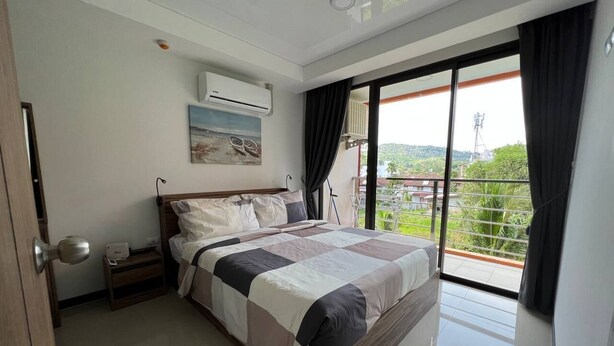 A bedroom with a large bed, a nightstand, an air conditioning unit, and a view of greenery through a balcony. - Title by Villacarte (Tambon Rawai)