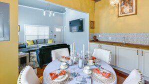 Un comedor con una mesa redonda dispuesta para cuatro personas, rodeada de sillas blancas. Se observa un sofá y un televisor al fondo.