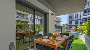 Een balkon met een houten tafel gedekt voor twee personen, omringd door rieten stoelen. Door de glazen deuren is een eethoek met stoelen en een tafel zichtbaar.