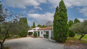 Une villa blanche, avec un toit recouvert de tuiles, entourée de verdure et avec une montagne en arrière-plan.