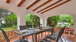 Un espace de restauration en plein air avec une longue table dressée pour quatre personnes, entourée de chaises, sous un plafond en bois.