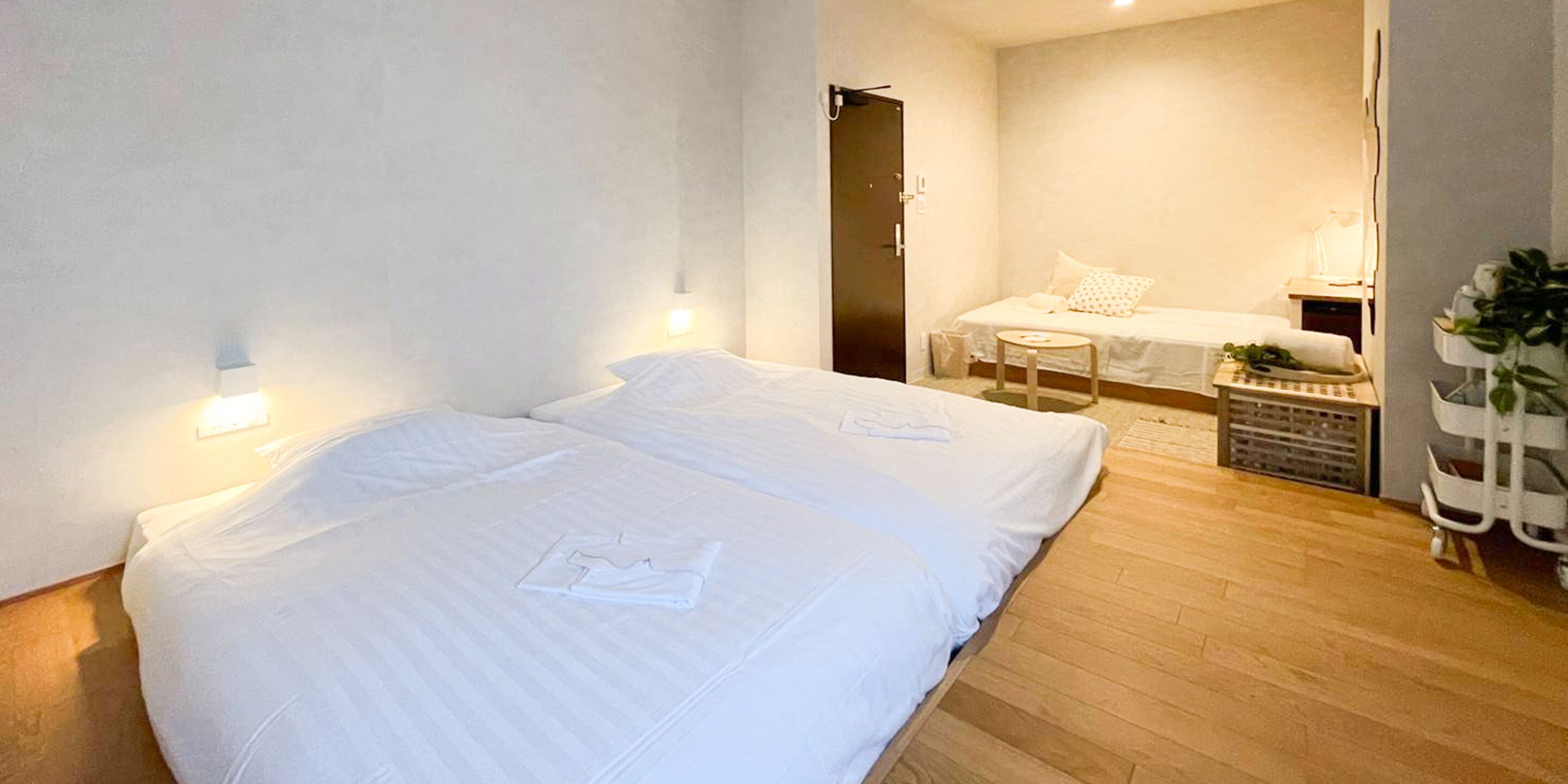 A hotel room with two beds, a small table, a plant, and a door leading to another room.