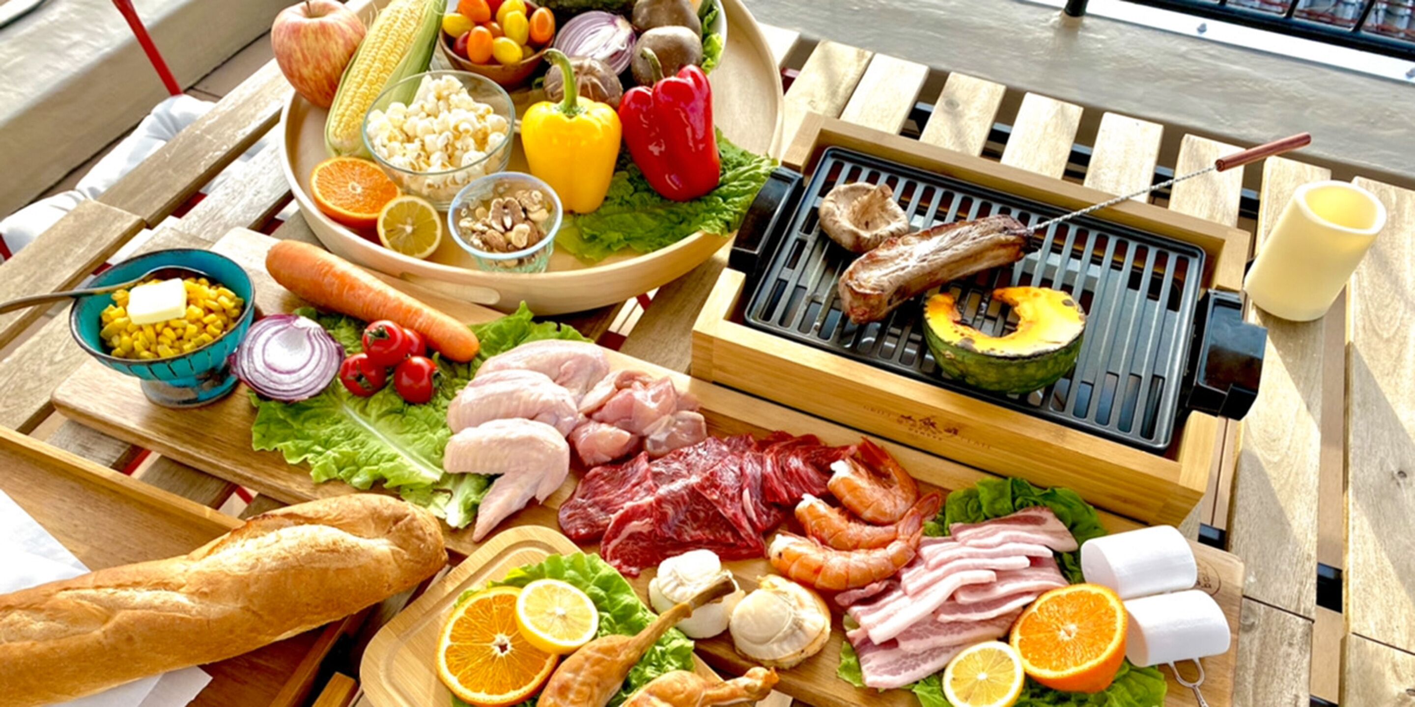 A variety of meats, vegetables, and fruits on a wooden table, with a grill and a bottle of water.
