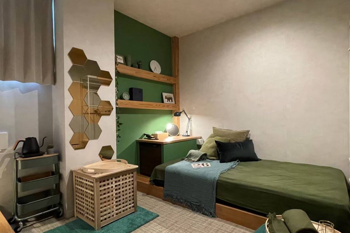 A compact bedroom with a bed, bedside table, lamp, and a wall-mounted shelf with various items.
