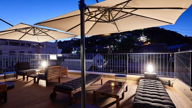 An outdoor seating area with wooden furniture, large umbrellas, and a view of a traditional building in the background. - Mange Tak Resort Onomichi (Onomichi)