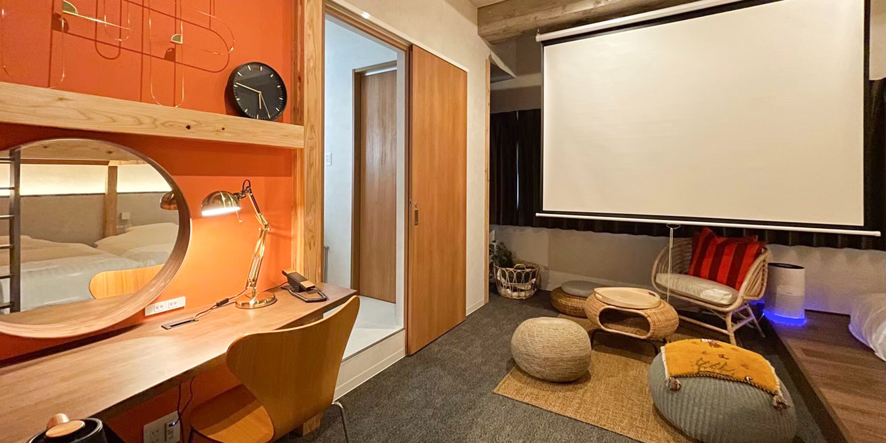 Ein modernes Hotelzimmer mit Schreibtisch aus Holz, einem großen Spiegel, einer runden Lampe, einer Projektionsleinwand und einer gemütlichen Sitzecke mit Kissen und einem kleinen Tisch.