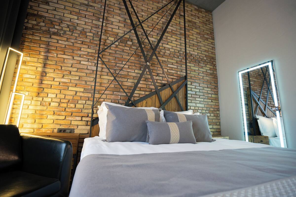 A modern hotel room with a brick wall, a bed with grey bedding, and a black leather chair.
