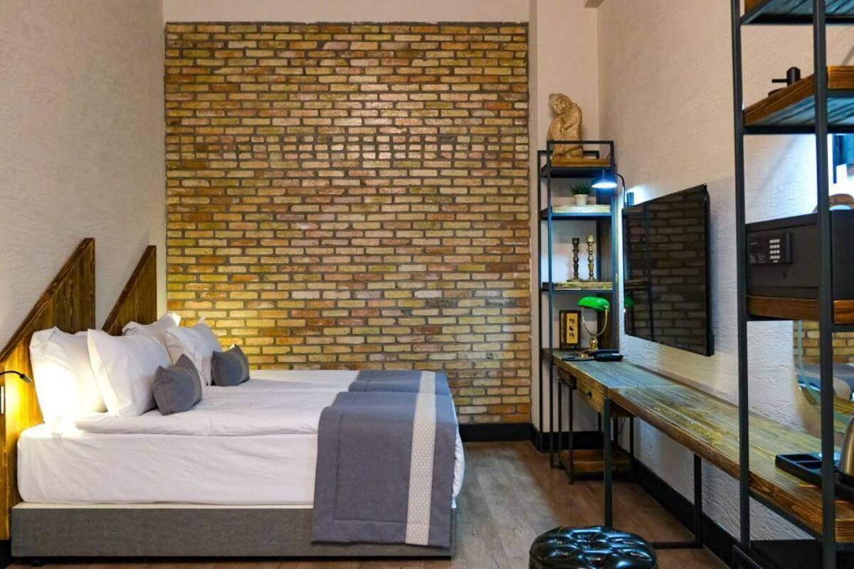 A bedroom with a brick wall, a bed with pillows, a bedside table with a lamp, and a shelf with decorative items.