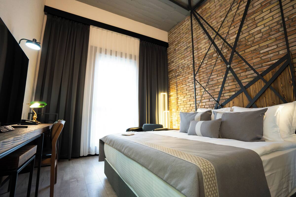 A hotel room with a large bed, a desk with a lamp, and a brick wall.