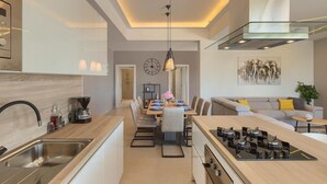Eine moderne Küche mit einer zentralen Kochinsel, einem Essbereich mit rundem Tisch und einer Wohnfläche inklusive Sofa und Couchtisch.