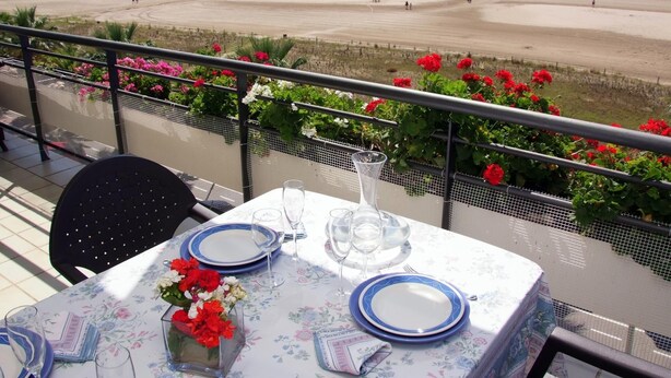 Ein Tisch ist mit Tellern, Gläsern und einer Blumenvase gedeckt, mit Blick auf einen Strand mit Menschen und einem Steg.