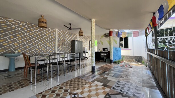 A modern dining area with a geometric patterned floor, a central dining table with chairs, a wall-mounted fan, and a ceiling fan. - Green Reed Homestay (Bokakhat)