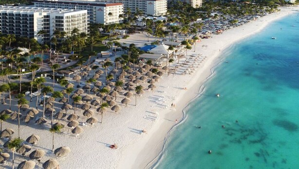 Ein Strandresort mit mehreren Hotelgebäuden, einem Sandstrand und strohgedeckten Sonnenschirmen.