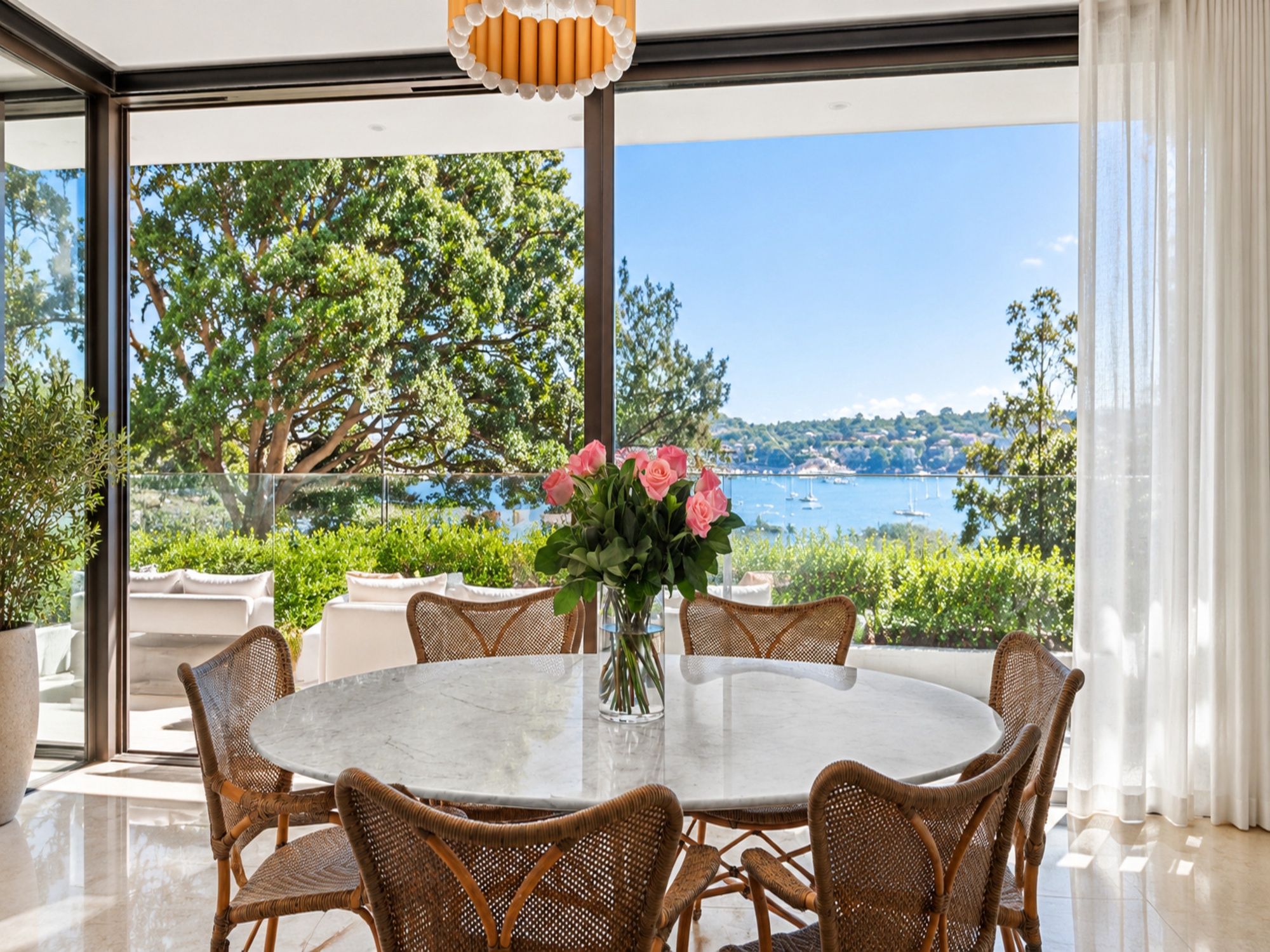 A round table with wicker chairs and a vase of flowers, set in a room with large windows overlooking a body of water and greenery.
