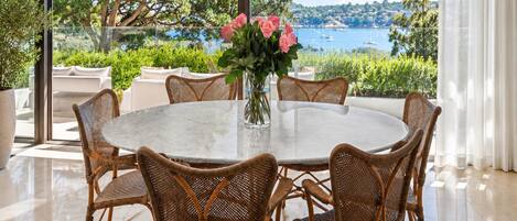 A round table with wicker chairs and a vase of flowers, set in a room with large windows overlooking a body of water and greenery.