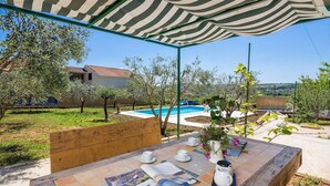 Un área junto a la piscina con una mesa de madera preparada para dos personas, un dosel rayado en la parte superior y vistas a una casa y árboles al fondo.