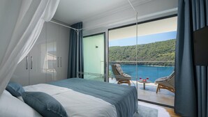 A bedroom with a bed, a balcony with wicker chairs, and a scenic view of a lake and forest.