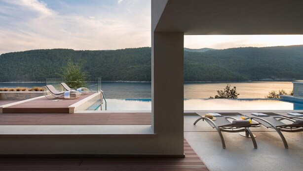 A modern outdoor pool area with lounge chairs and a view of a lake and forested hills.