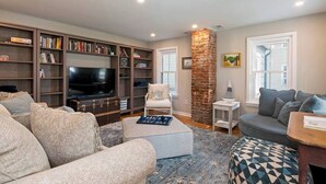A living room with a large flat-screen TV, a bookshelf, a white armchair, and a grey sofa. - Pet-Friendly Tilghman Island Coastal Retreat (Tilghman Island)