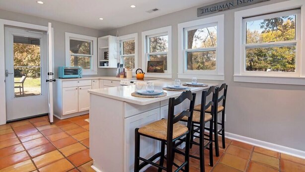 A kitchen with white cabinets, a large island with bar stools, and a view of trees through the windows. - Pet-Friendly Tilghman Island Coastal Retreat (Tilghman Island)