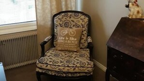 Una habitación de esquina con un sillón estampado, una mesita con un peluche y una ventana con persianas.