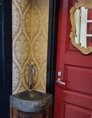 Une porte rouge ornée d’un cadre doré, un mur recouvert d’un papier peint à motifs et un évier en pierre avec un robinet.