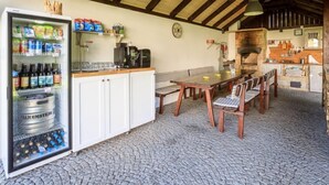 A rustic kitchen with a stone floor, wooden beams, a refrigerator stocked with beverages, a coffee machine, and a dining area with wooden chairs and a table.
