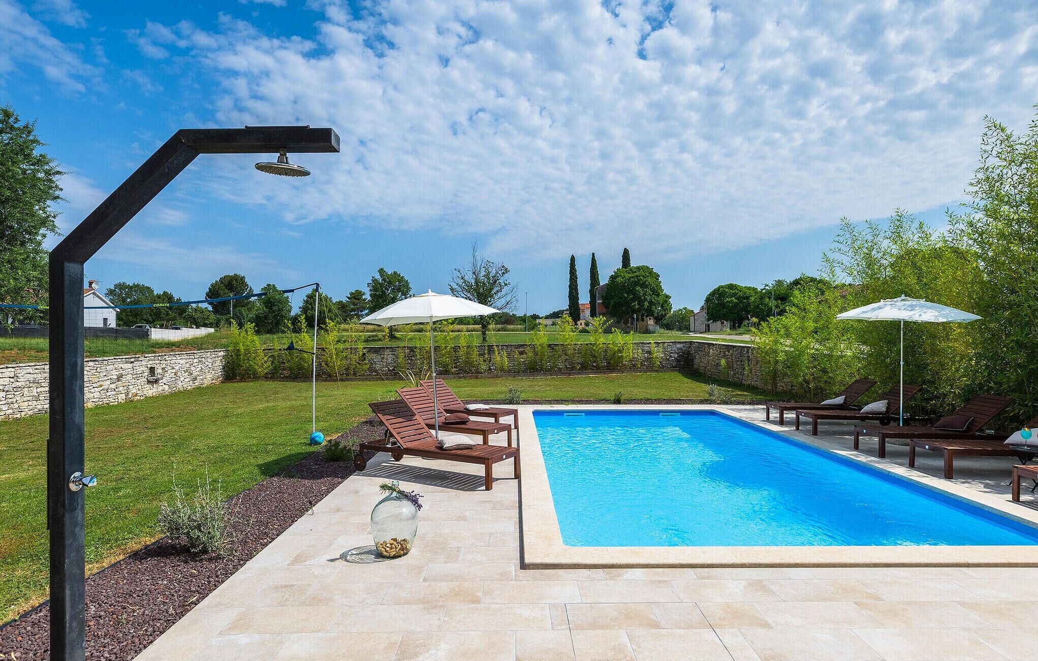 A swimming pool with lounge chairs and umbrellas, surrounded by a stone wall and greenery.