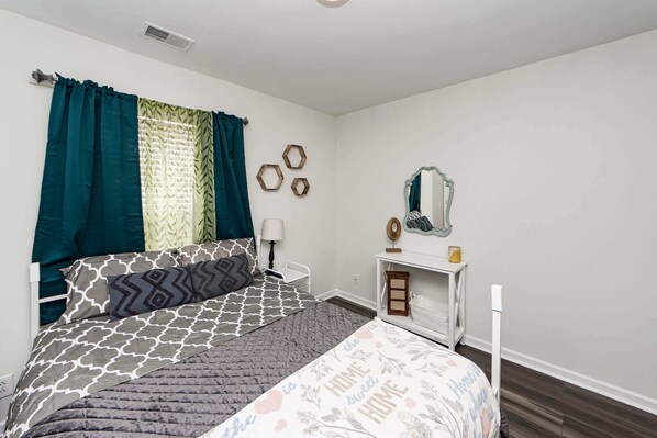 A bedroom with a bed, a bedside table, a mirror, and decorative wall art. - The Oasis at Saratoga (North Charleston)