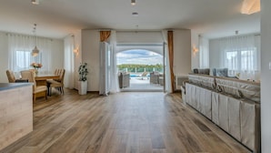 Ampla sala de estar com piso de madeira, conjunto de jantar, sofá e vista para uma piscina e vegetação.