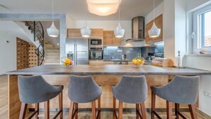 Cozinha moderna com ilha central, quatro banquetas, eletrodomésticos de aço inoxidável e iluminação pendente.