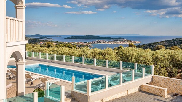 Piscina com guarda-corpo de vidro, rodeada por vegetação e com vista para a paisagem costeira.