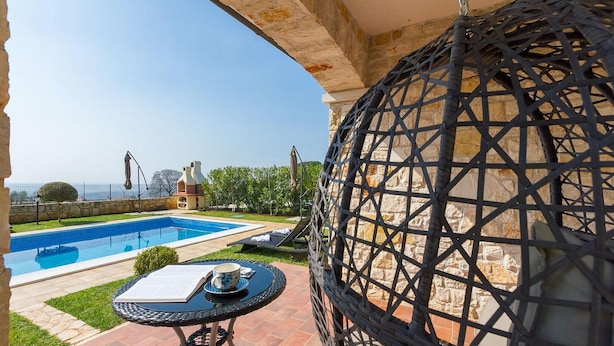 A pool area with a view of the sea, a round table with a book and a cup, and a decorative black spherical object.
