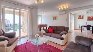 A spacious living room with a brown sofa, a glass coffee table, and a red rug. There is a dining area with a table and chairs, and a view of a pool through large windows.