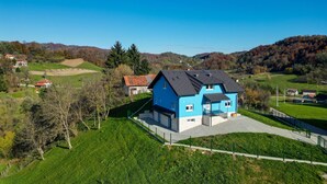 Una casa azul con entrada pavimentada y patio cercado, rodeada de campos verdes y árboles.