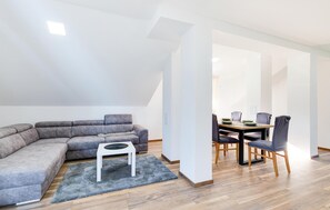 Un salon moderne avec un canapé d’angle gris, une petite table d’appoint blanche et un coin repas comprenant une table et des chaises en bois.