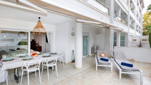 A spacious outdoor dining area with a white table and chairs, a hanging light fixture, and a view of the building's exterior. - Cosy apartment for 6 guests with A/C, pool, WIFI, TV and terrace (Castell-platja D'aro)