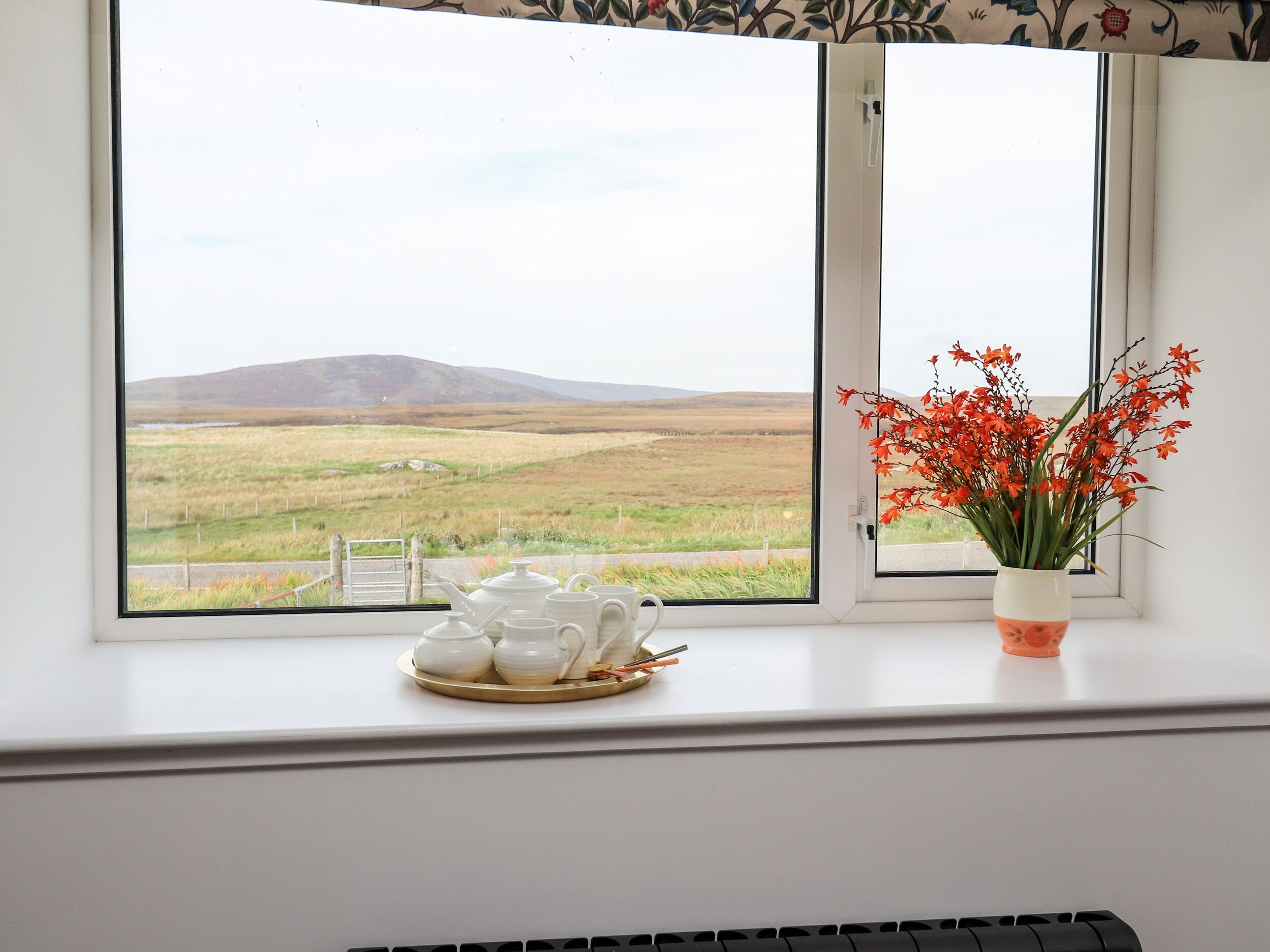 Ein Fenster mit Blick auf ein grasbewachsenes Feld und Hügel, ein weißes Tee-Set und eine Vase mit roten Blumen auf einem Fensterbrett.