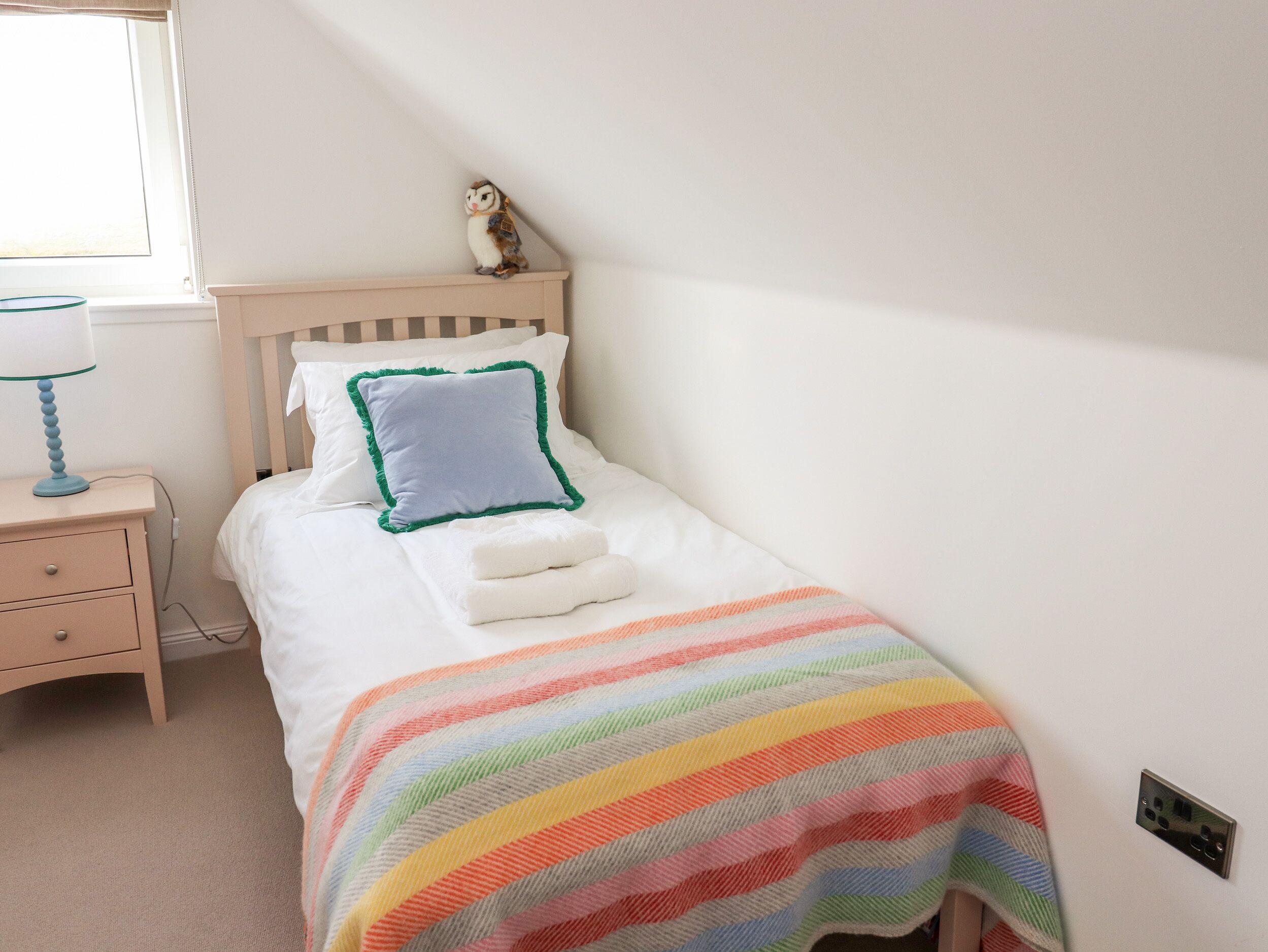 Ein ordentlich bezogenes Bett mit einer Decke im Regenbogenstreifen, ein kleiner Nachttisch mit Lampe sowie ein an der Wand befestigtes Regal mit einer Figur.